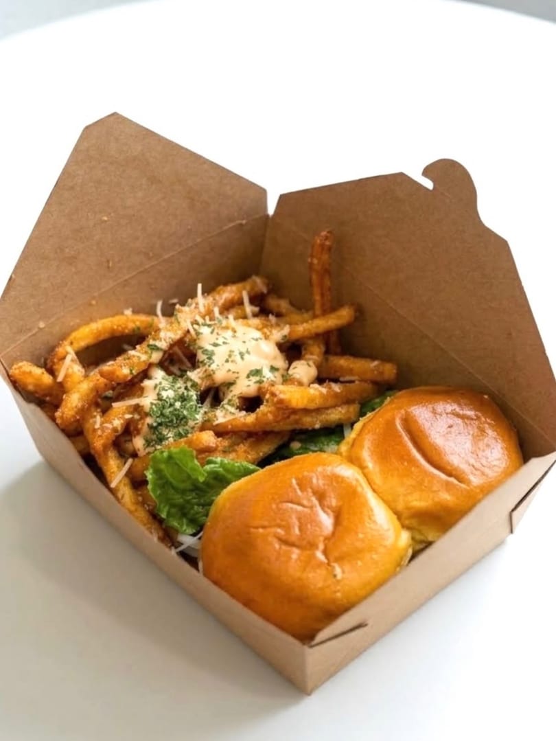 Two sliders and seasoned curly fries with sauce and herbs in a brown takeout box.