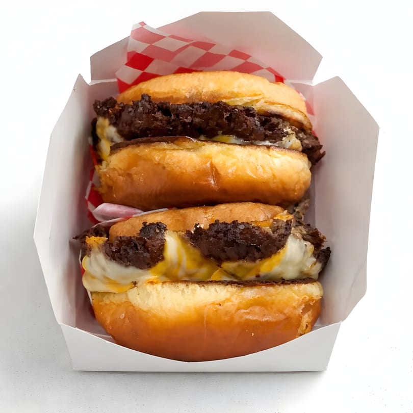 Two cheeseburger sliders with melted cheese and beef patties on glazed donut buns in a white box.